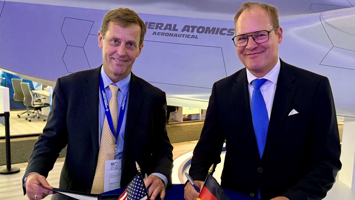 Two men wearing suits smiling at the camera as they sign documents, with small versions of the American flag and German flag on the table.