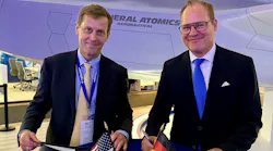 Two men wearing suits smiling at the camera as they sign documents, with small versions of the American flag and German flag on the table.