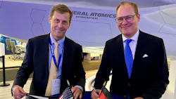 Two men wearing suits smiling at the camera as they sign documents, with small versions of the American flag and German flag on the table. Two men wearing suits smiling at the camera as they sign documents, with small versions of the American flag and German flag on the table.