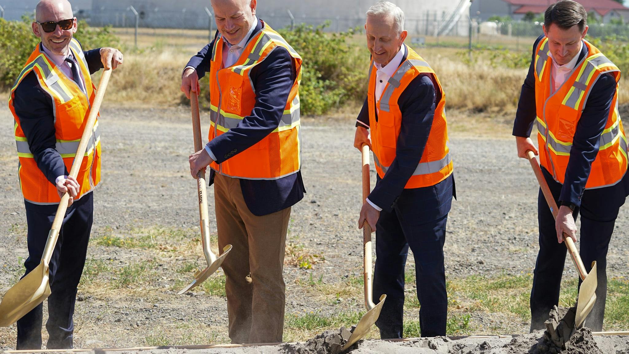 Four men wearing business attire and bright orange vests digging shovels into a pile of dirt