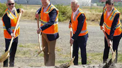 Four men wearing business attire and bright orange vests digging shovels into a pile of dirt