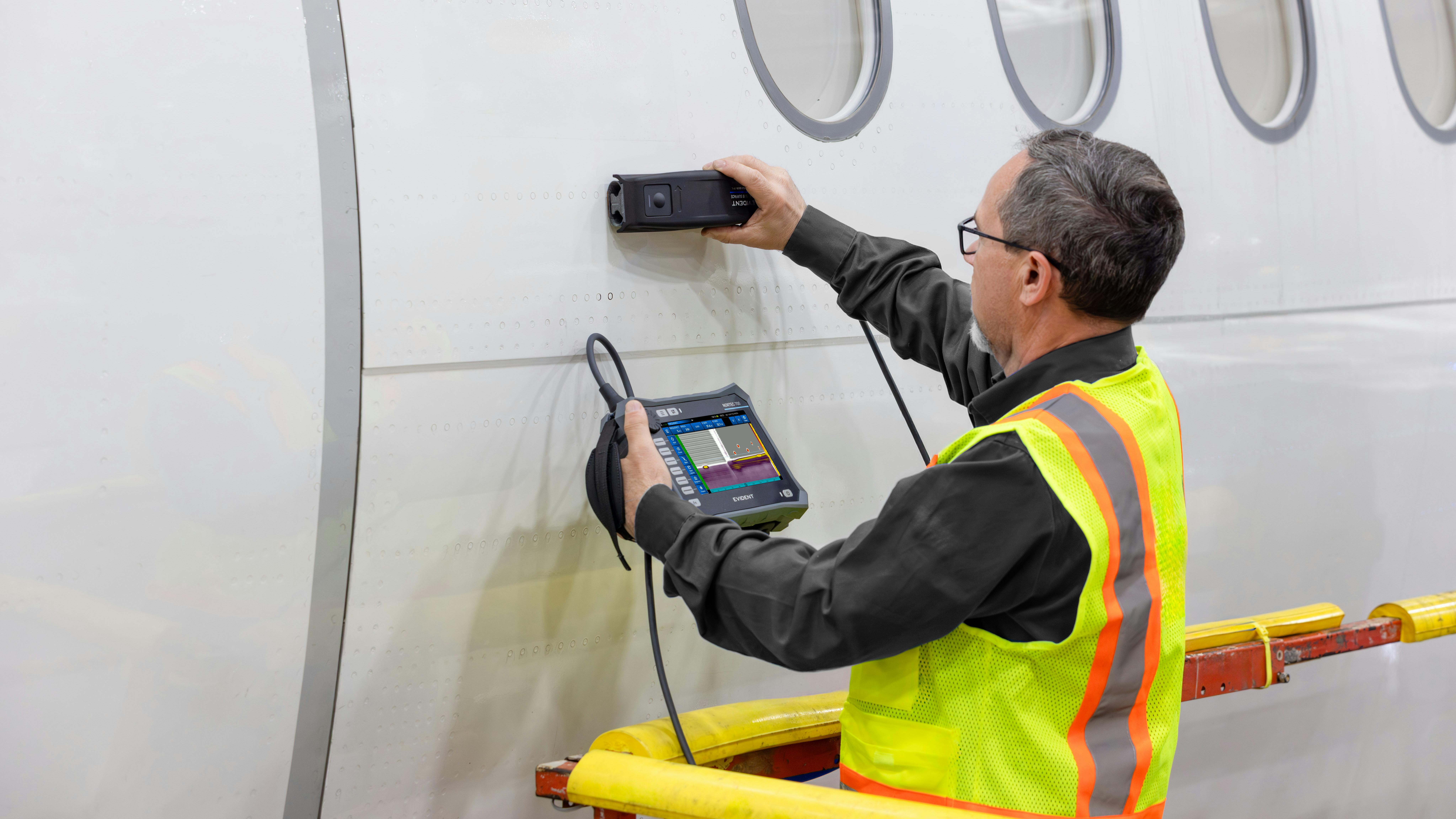 A man wearing a high-visibility vest holding a tool up to the side of an airplane while holding a screen