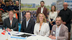 Three men and one woman wearing business attire and sitting at a table for a document signing, with people observing from behind them Three men and one woman wearing business attire and sitting at a table for a document signing, with people observing from behind them