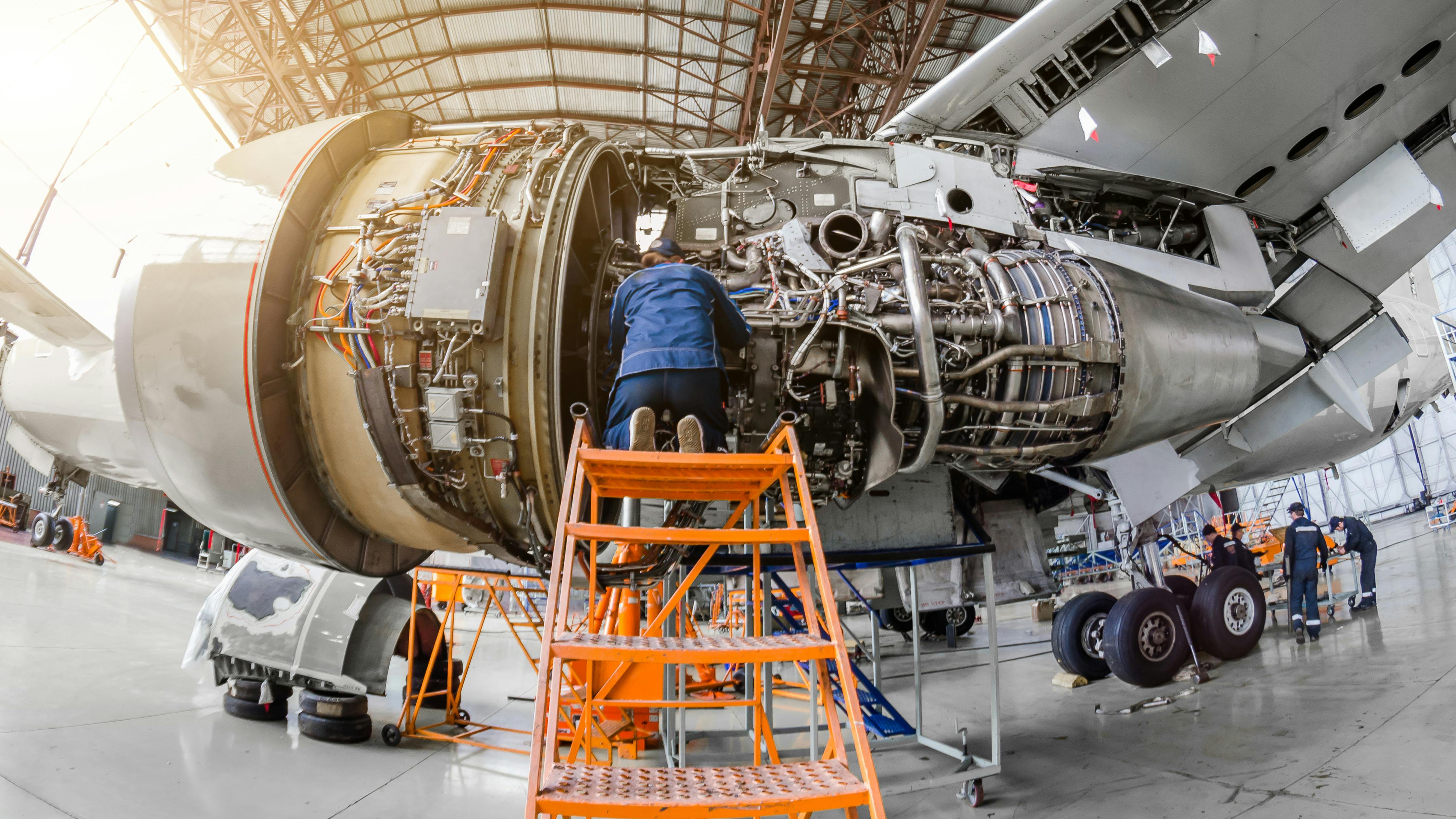 A technician at the top of an orange work platform with stairs, working on an opened-up airp[ane engine