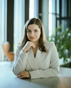 A woman wearing business attire sitting at a table and smiling at the camera, resting her chin on her hand A woman wearing business attire sitting at a table and smiling at the camera, resting her chin on her hand