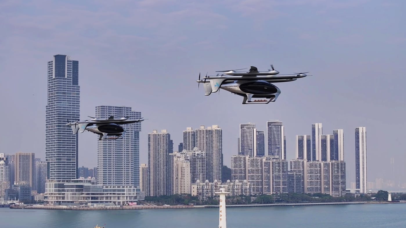 Two drone-like aircraft hovering in the air in front of a city skyline and body of water
