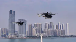 Two drone-like aircraft hovering in the air in front of a city skyline and body of water