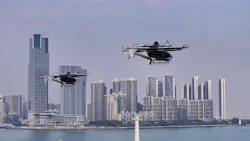 Two drone-like aircraft hovering in the air in front of a city skyline and body of water Two drone-like aircraft hovering in the air in front of a city skyline and body of water
