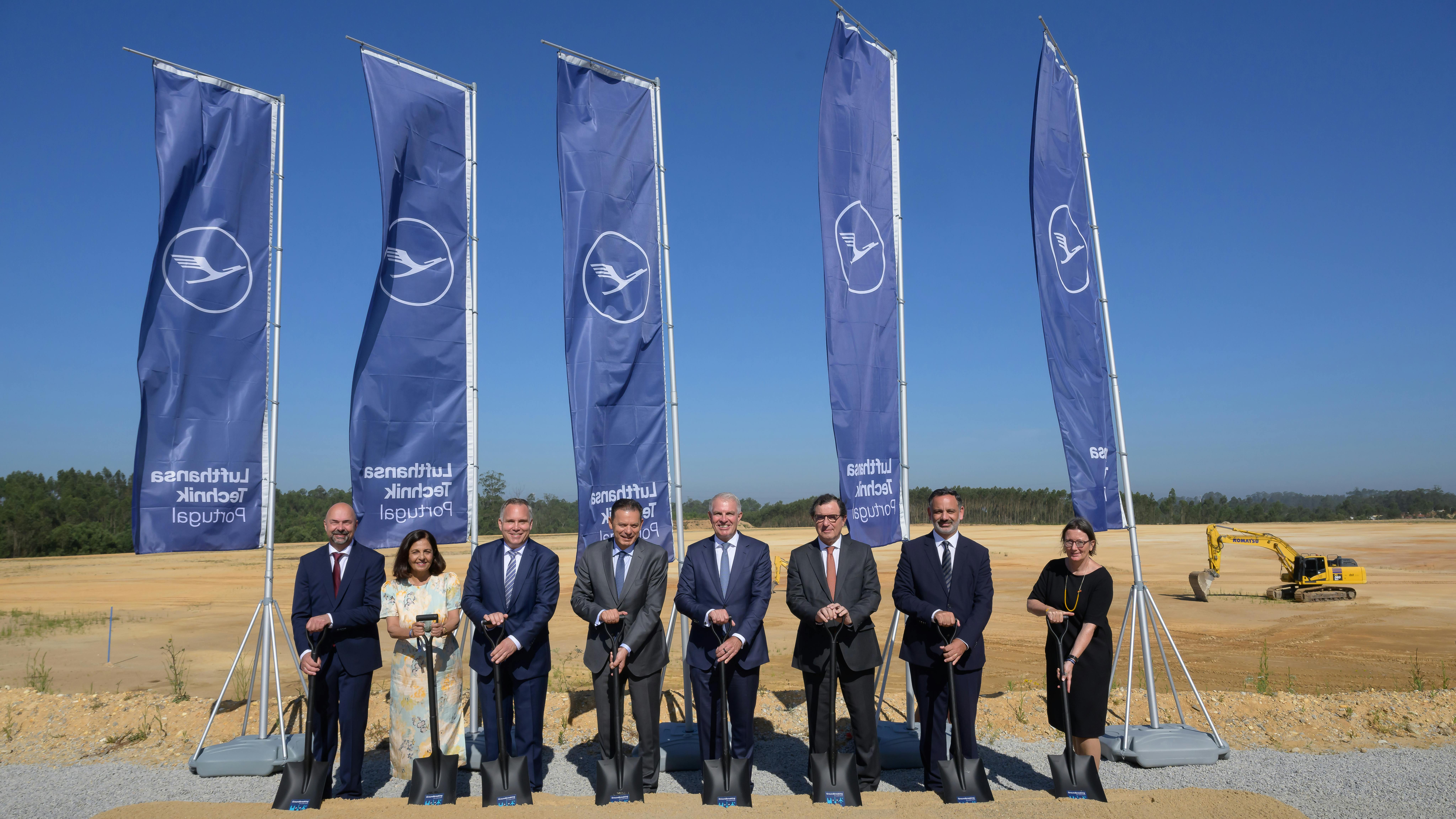 A group of professionals wearing business attire standing at a construction site, digging shovels into the ground