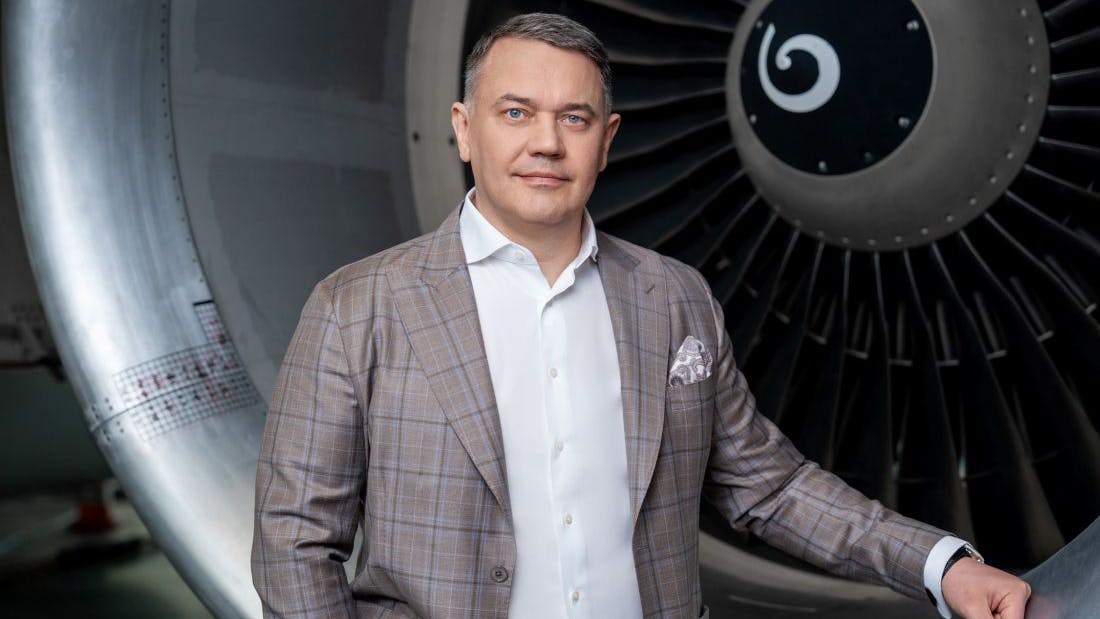 A man wearing a suit looking sternly into the camera while standing in front of an airplane engine