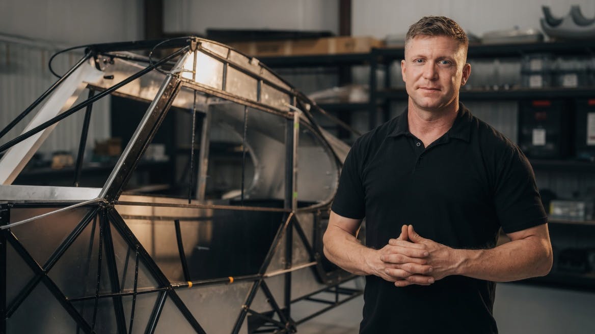 A man wearing a black polo smiling at the camera while standing next to a deconstructed airplane