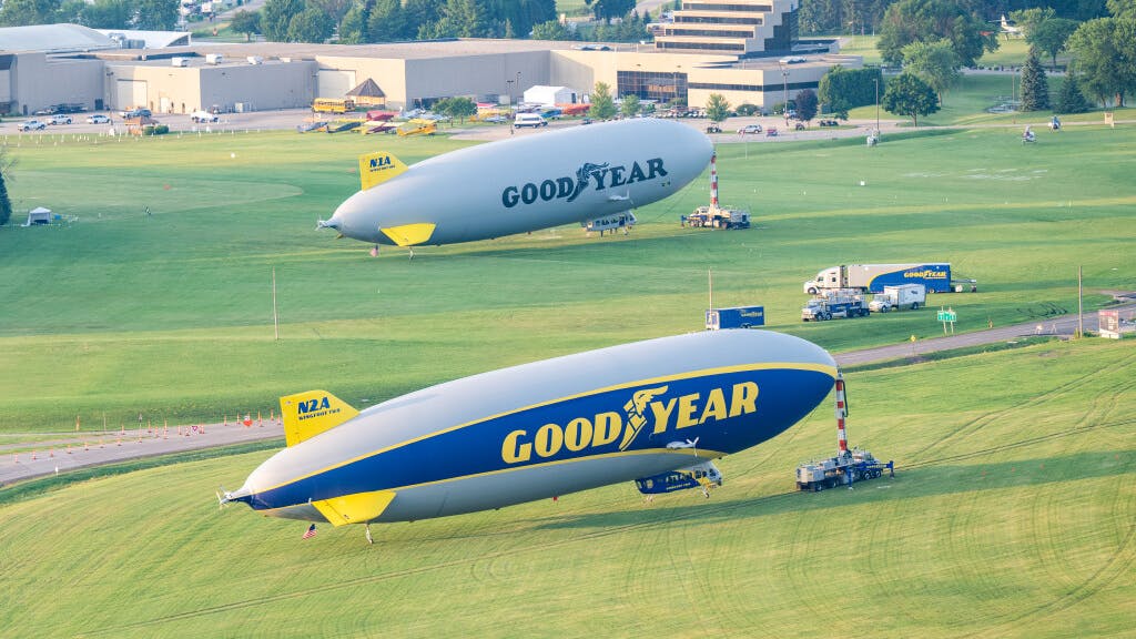 Two large blimps, one gray and one blue and yellow, parked on grass, both with the word 'GOODYEAR' printed on the side