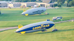 Two large blimps, one gray and one blue and yellow, parked on grass, both with the word 'GOODYEAR' printed on the side