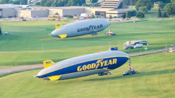 Two large blimps, one gray and one blue and yellow, parked on grass, both with the word 'GOODYEAR' printed on the side