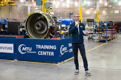 A man speaking into a microphone while standing in front of an airplane engine and a sign that reads: MTU Maintenance Training Center