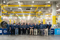 A group of people wearing dark blue shirts standing in a repair shop next to an airplane engine