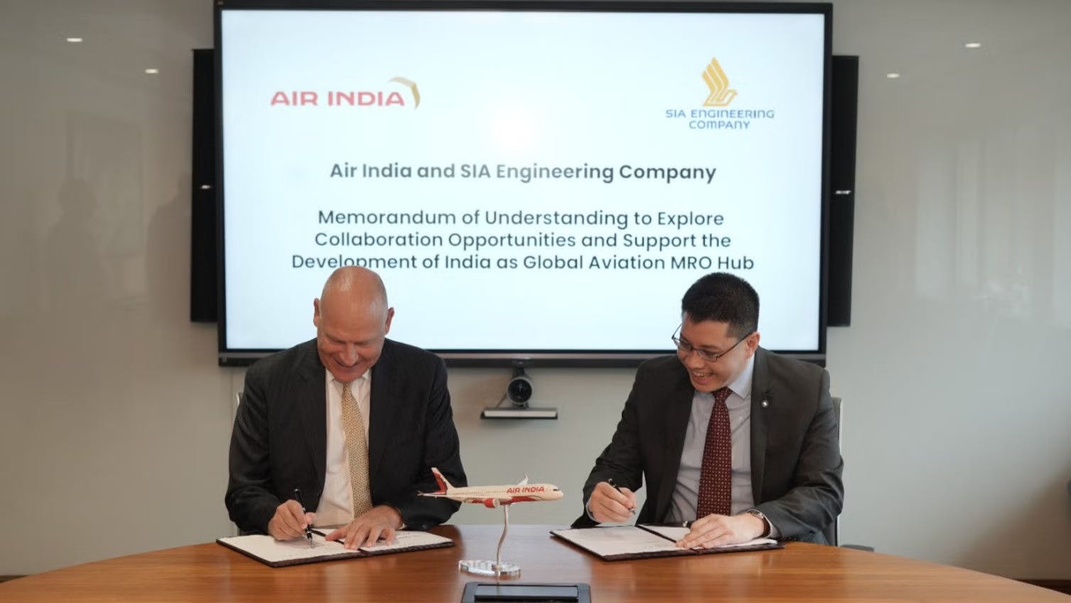 Two men wearing suits sitting at a table and signing pieces of paper, with text on a board behind them that reads: Air India and SIA Engineering Company Memorandum of Understanding to Explore Collaboration Opportunities and Support the Development of India as a Global Aviation MRO Hub