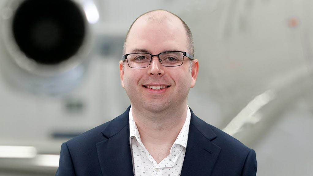 A close-up photograph of a man wearing a suit and glasses smiling at the camera