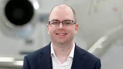 A close-up photograph of a man wearing a suit and glasses smiling at the camera