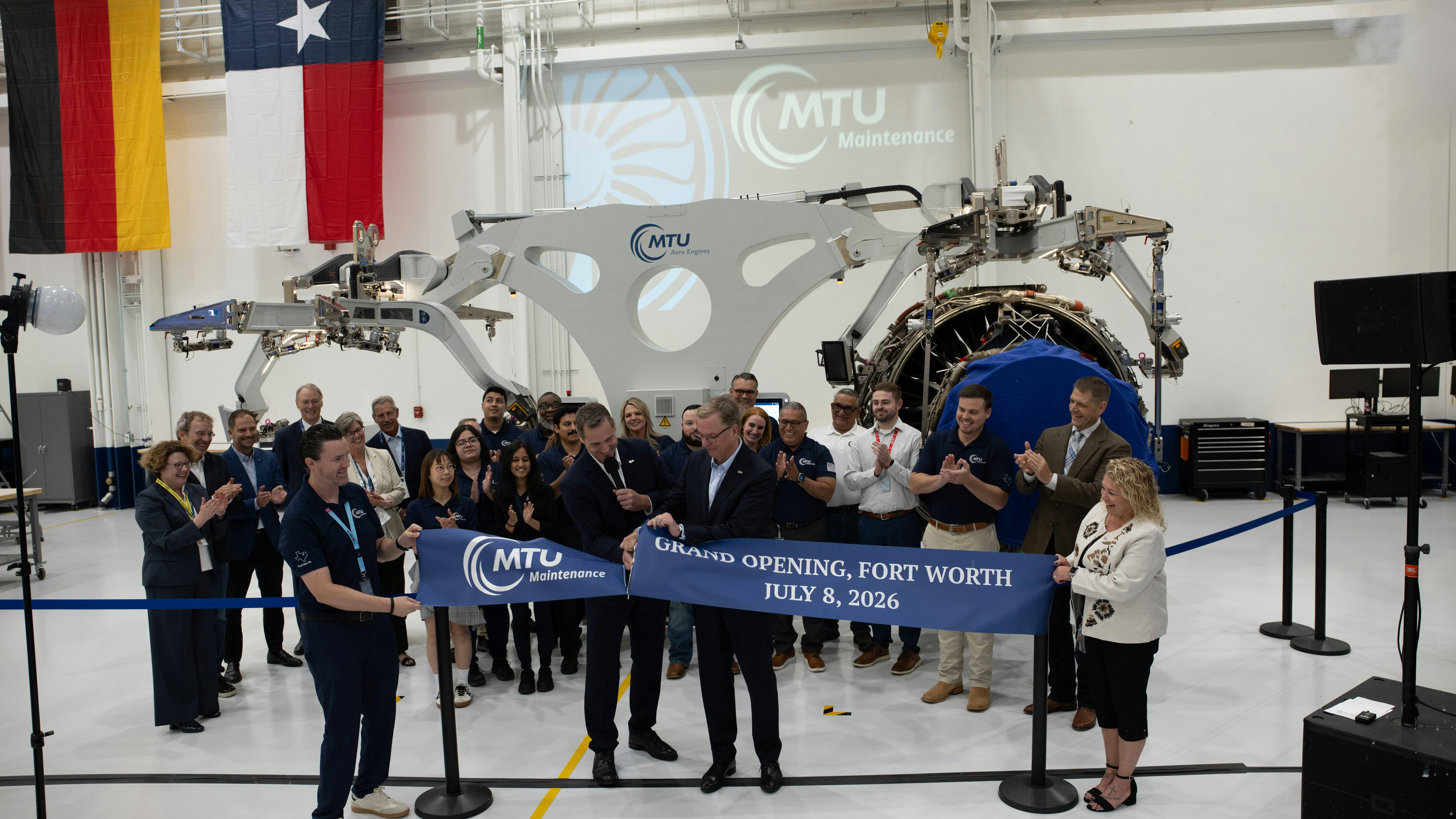 A crowd of people gathering around a red ribbon while someone cuts it, all standing inside an aircraft repair shop with a sign on the wall that says 'MTU Maintenance'