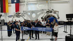 A crowd of people gathering around a red ribbon while someone cuts it, all standing inside an aircraft repair shop with a sign on the wall that says 'MTU Maintenance'