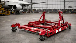 A red metal engine stand sitting on the floor in a repair shop with an airplane in the background