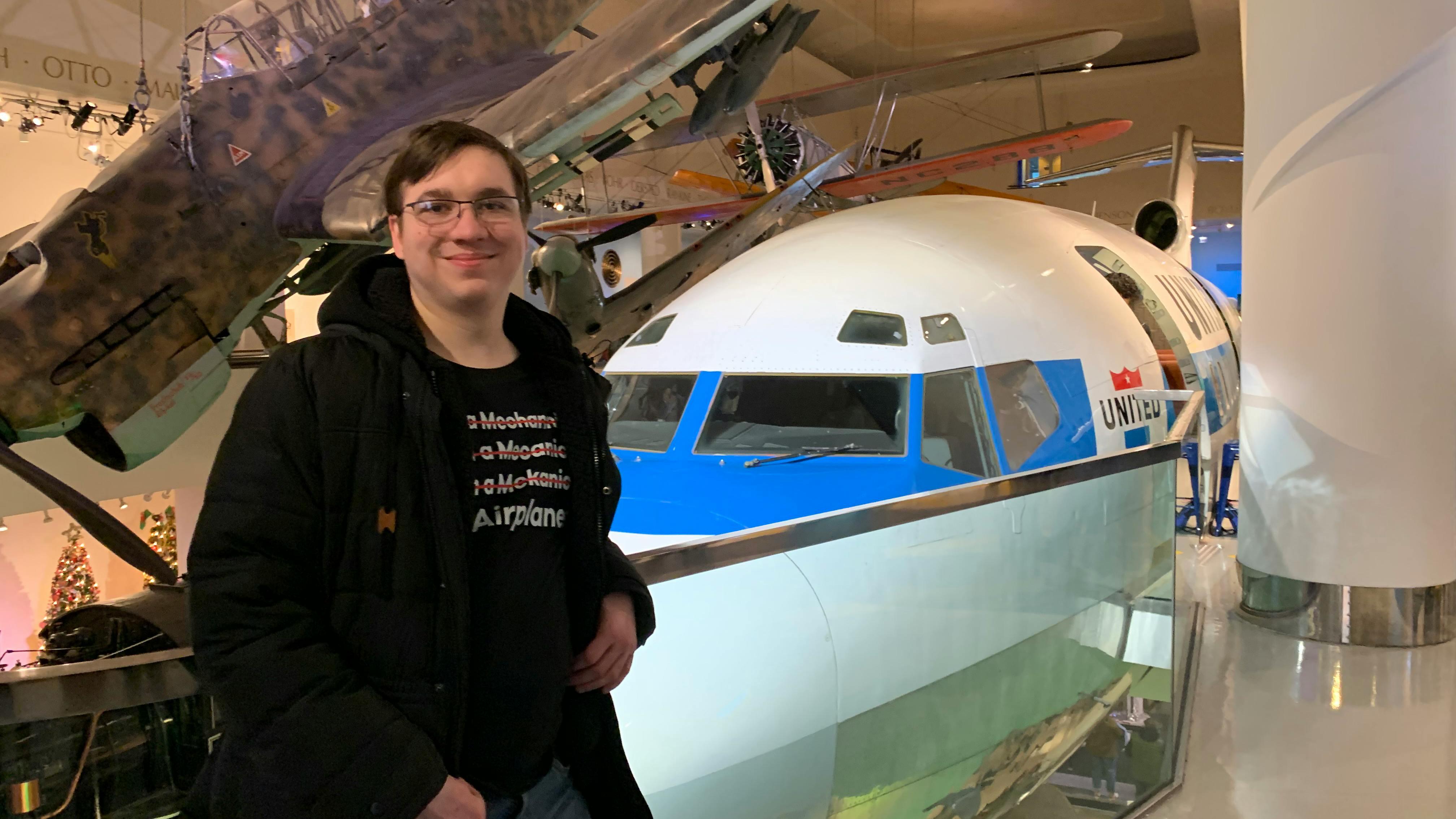 A man wearing a graphic tee and jacket standing in front of an airplane in a museum