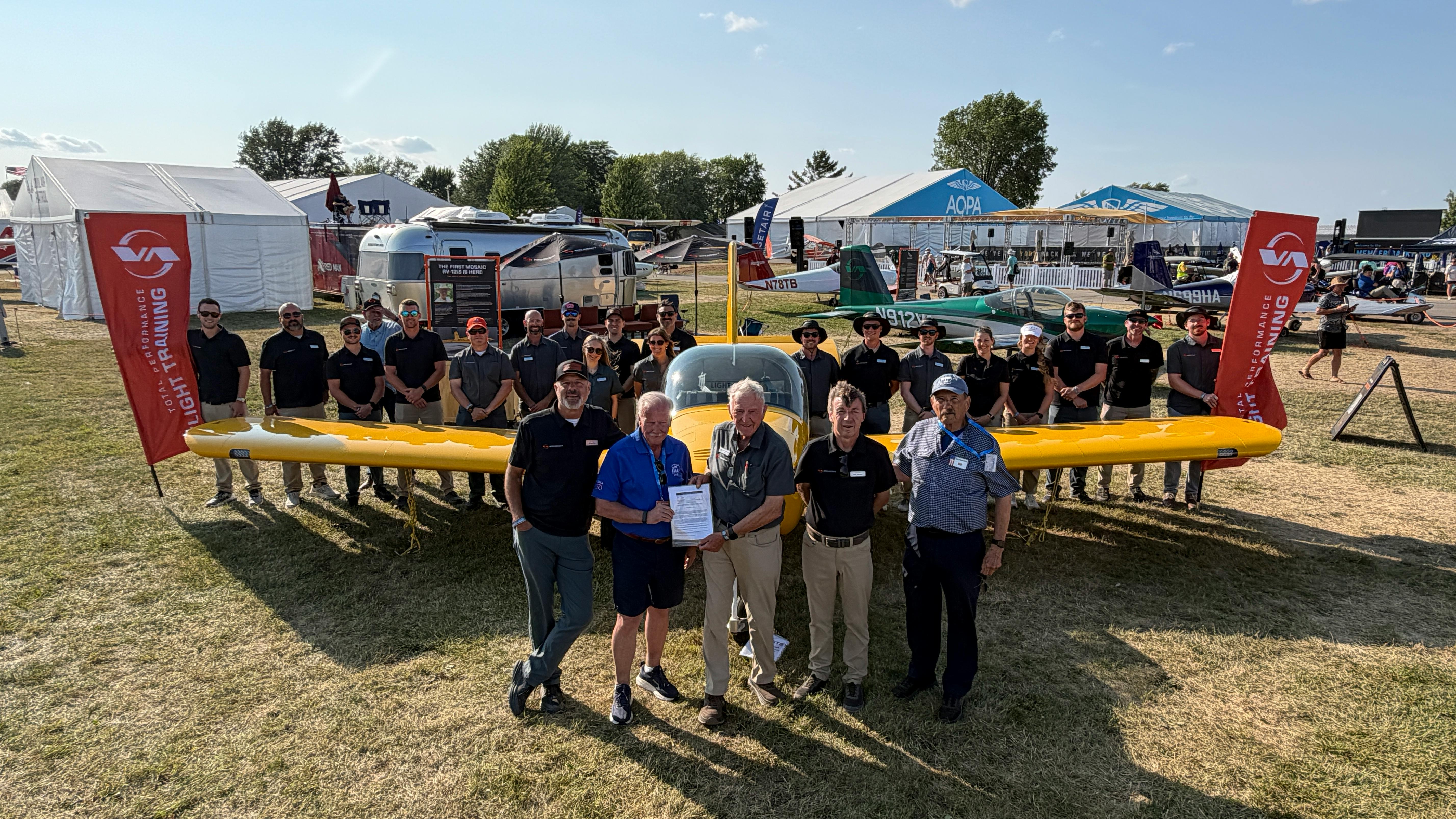 The Van's Aircraft team joines Van's Aircraft founder Dick VanGrunsven (front row, center) and EAA CEO/Chairman Jack Pelton (front row, second from left) on July 24, 2026, as the first MOSAIC-compliant RV-12 is delivered during EAA AirVenture Oshkosh 2026. Front row: Left to right: Mikael Via, Van&rsquo;s Aircraft CEO; Pelton; VanGrunsven; Rian Johnson, Van&rsquo;s Chief Engineer; and Designated Airworthiness Representative (DAR) Bill Kanino.