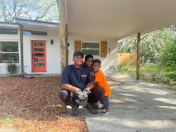 A man wearing a dark blue uniform crouching in front of a white house while two small children hug him