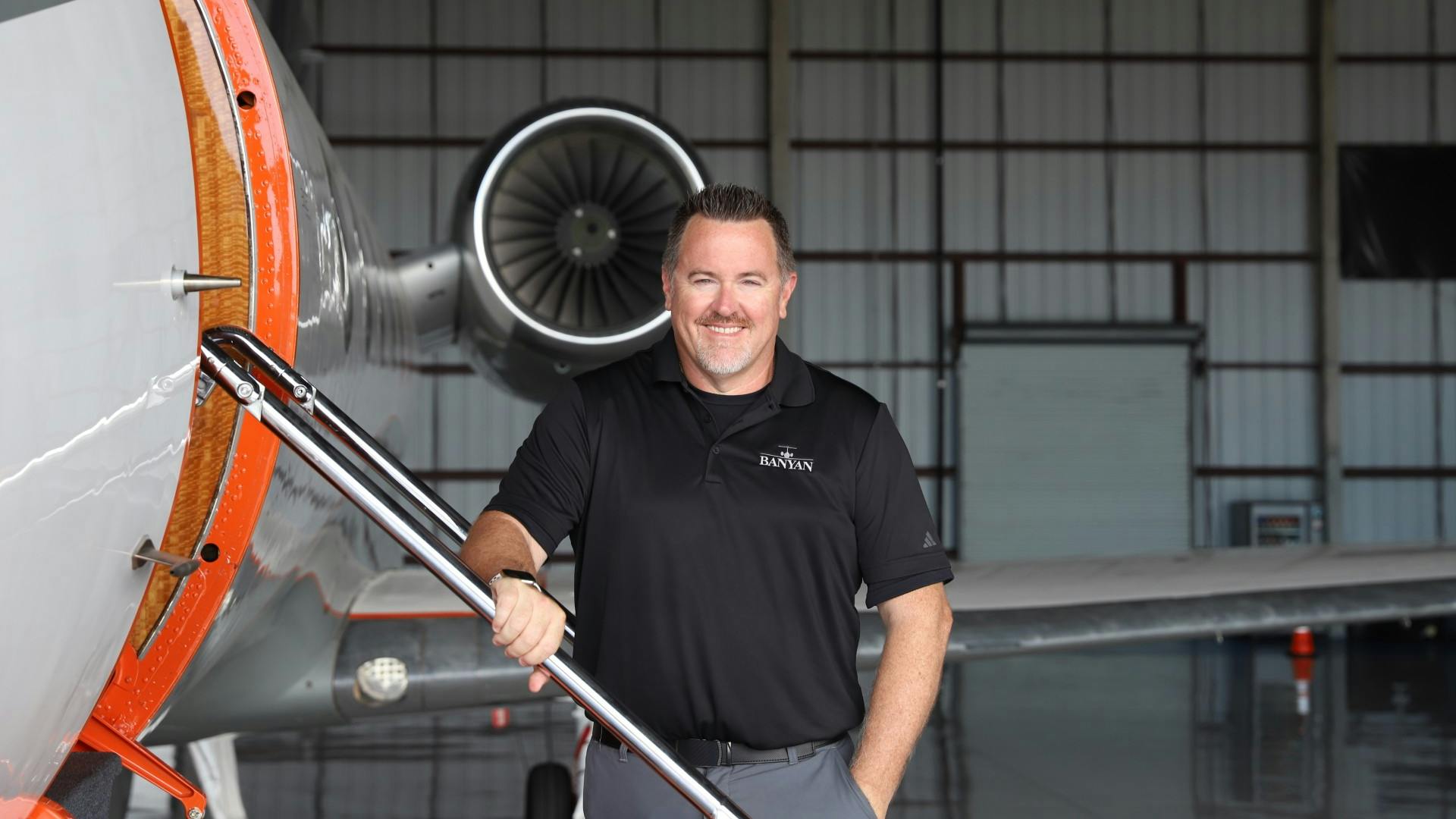 A man wearing a black polo shirt smiling at the camera while standing next to an airplane