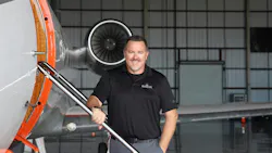 A man wearing a black polo shirt smiling at the camera while standing next to an airplane