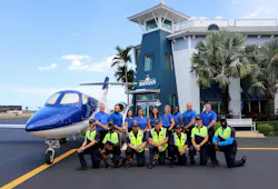 A crowd of people wearing blue polos with some in high-visibility vests standing near a small airplane in front of a building with a sign that says 'BANYAN'