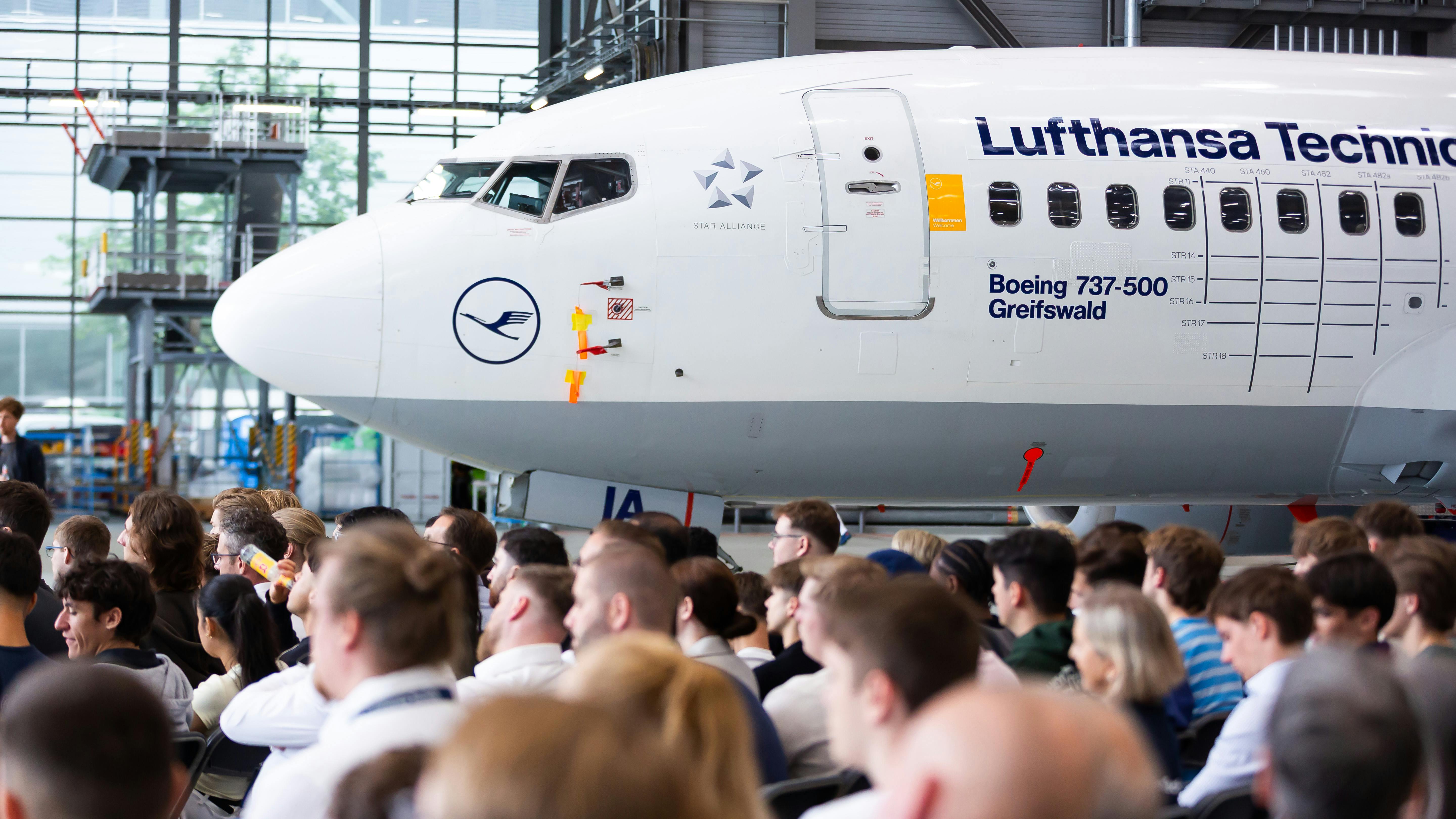 A large crowd of people standing inside an aircraft hangar with an airplane in the background labeled with text that reads: LUFTHANSA