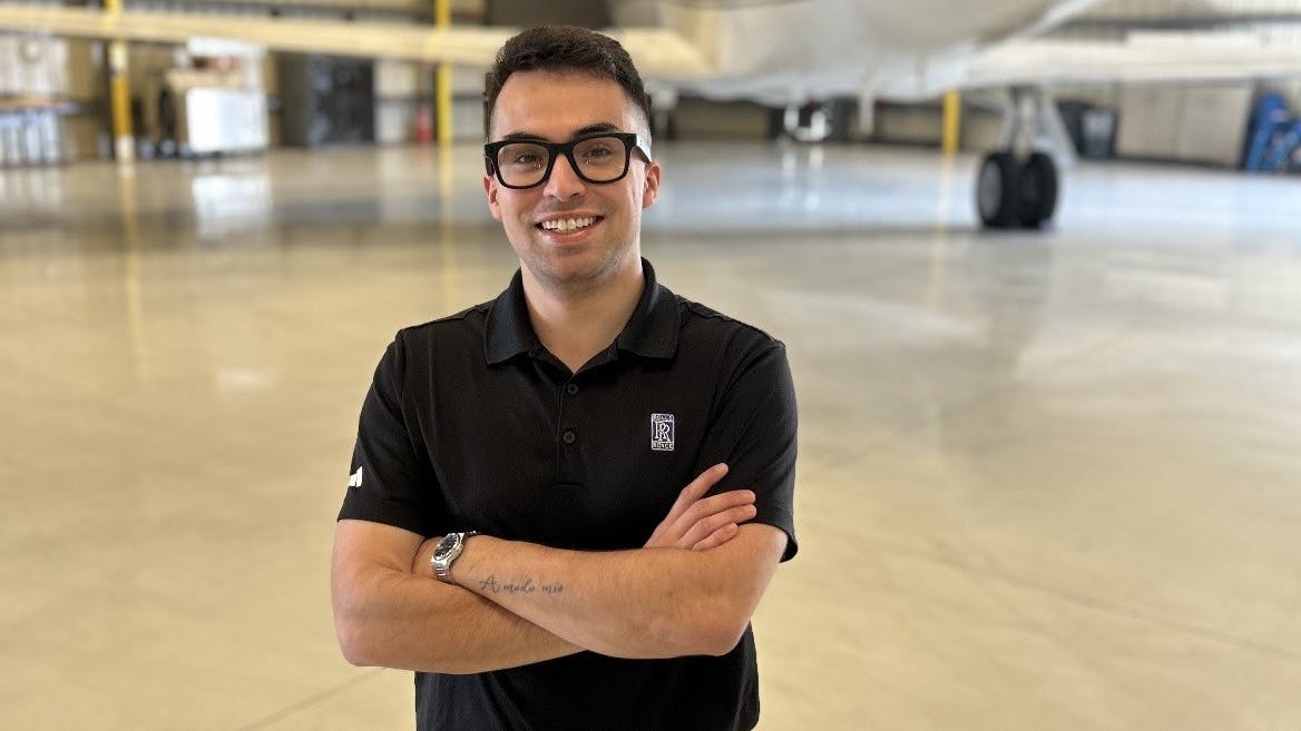 A man wearing glasses and a black polo crossing his arms and smiling at the camera while standing in front of an airplane