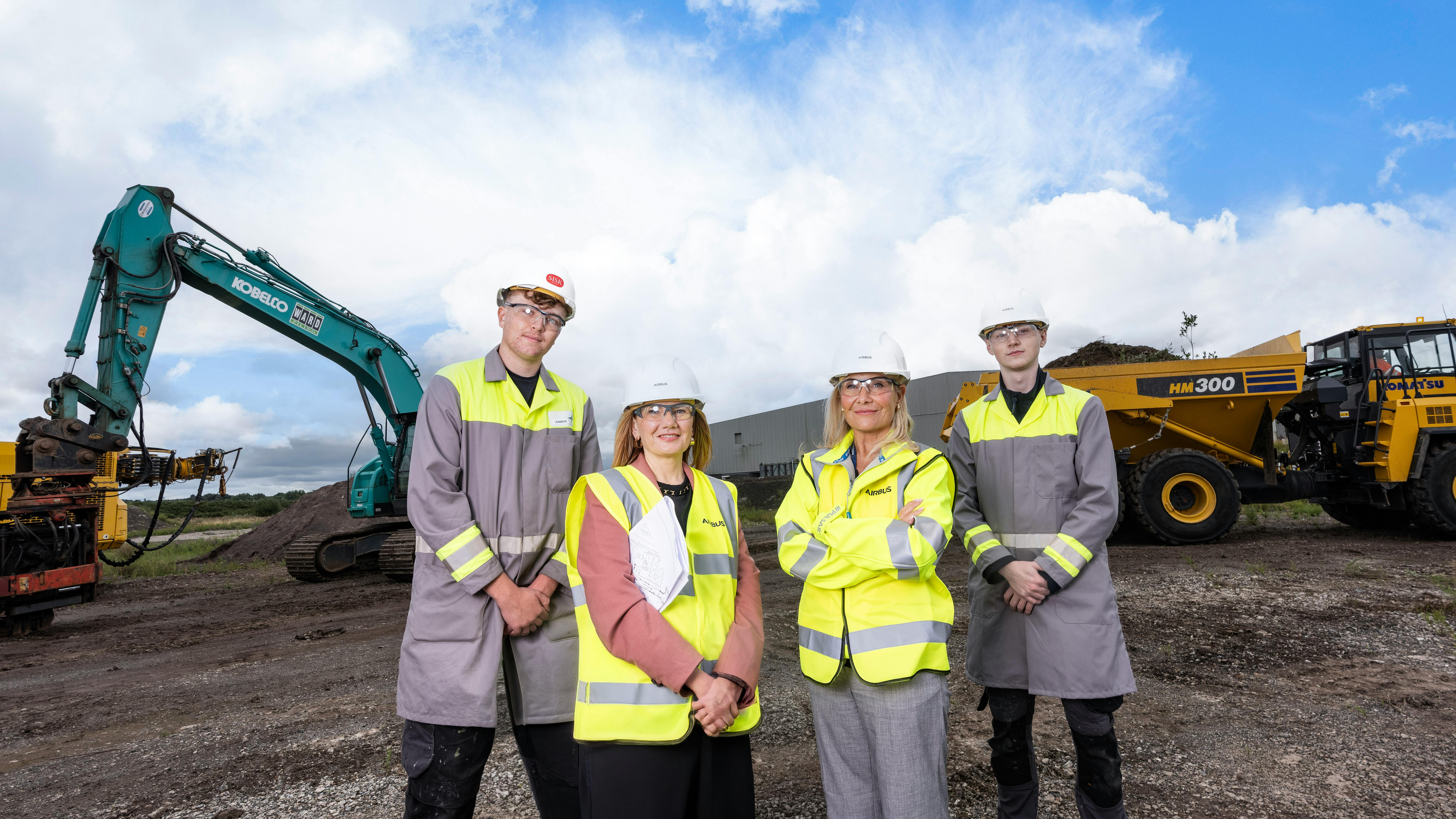 Two men and two women wearing white hard hats and high-visibility vests standing in front of a construction site