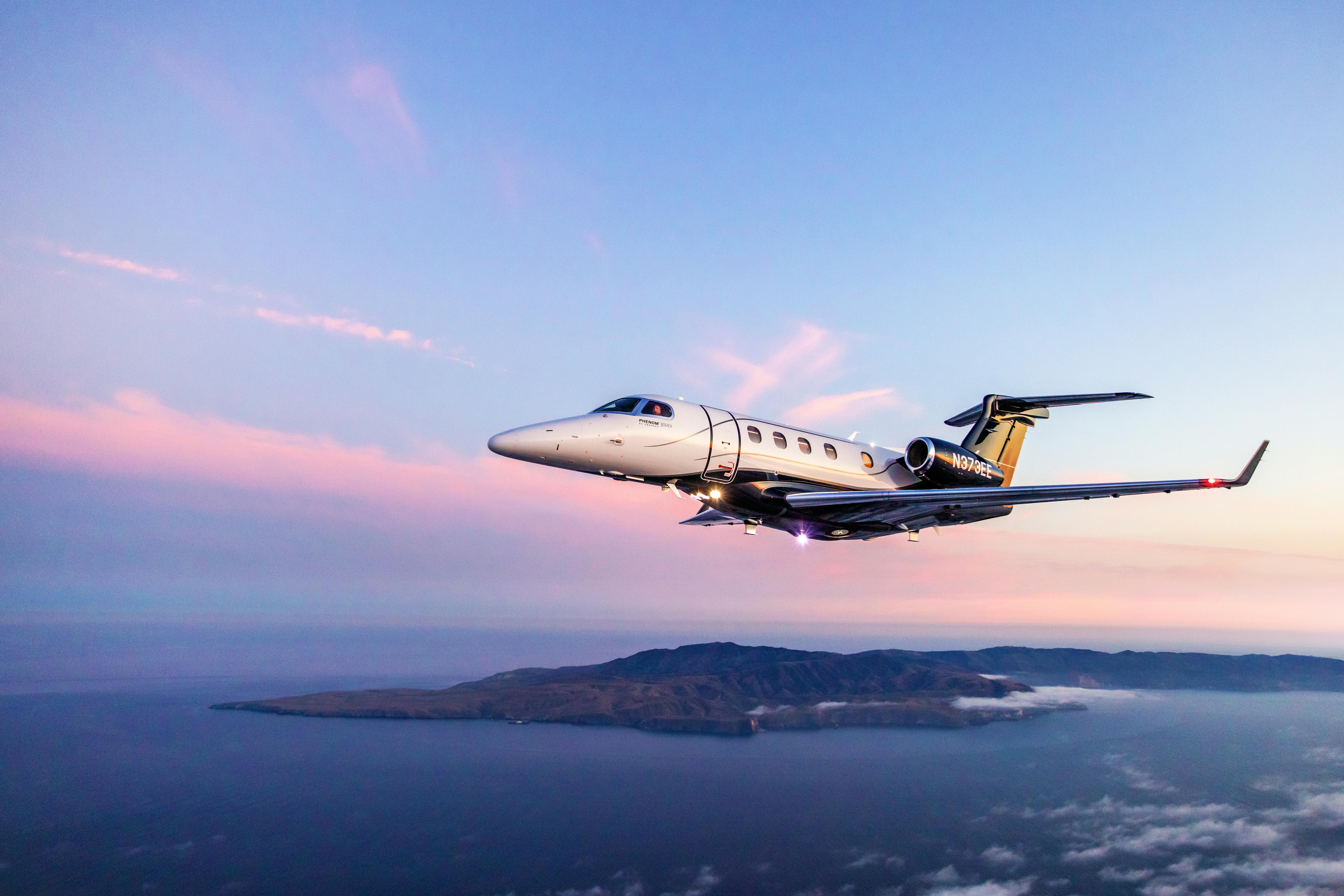 A gray aircraft flying over a body of water with an island and a sunset in the background