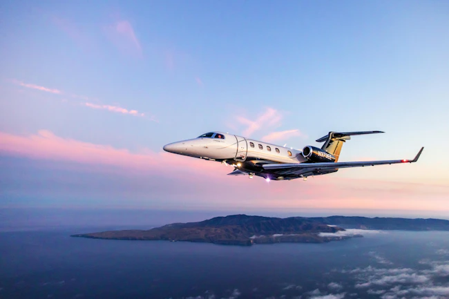 A gray aircraft flying over a body of water with an island and a sunset in the background