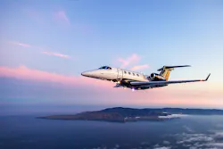 A gray aircraft flying over a body of water with an island and a sunset in the background