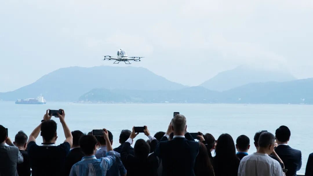 A large crowd of people watching and taking photos of an aircraft in the air over a body of water