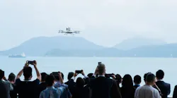 A large crowd of people watching and taking photos of an aircraft in the air over a body of water