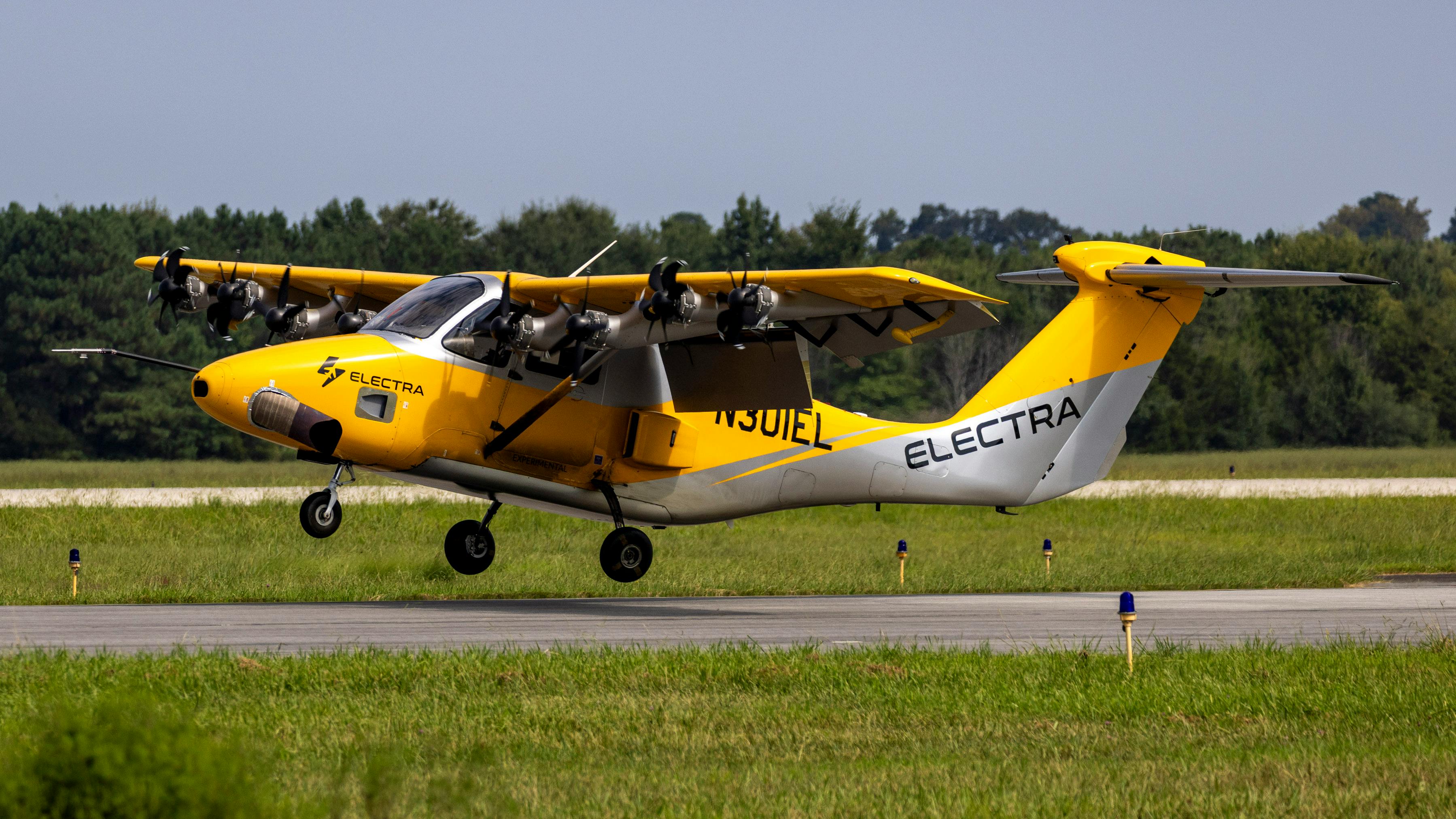 A small yellow aircraft with 'ELECTRA' printed on the side flying near the ground on a runway