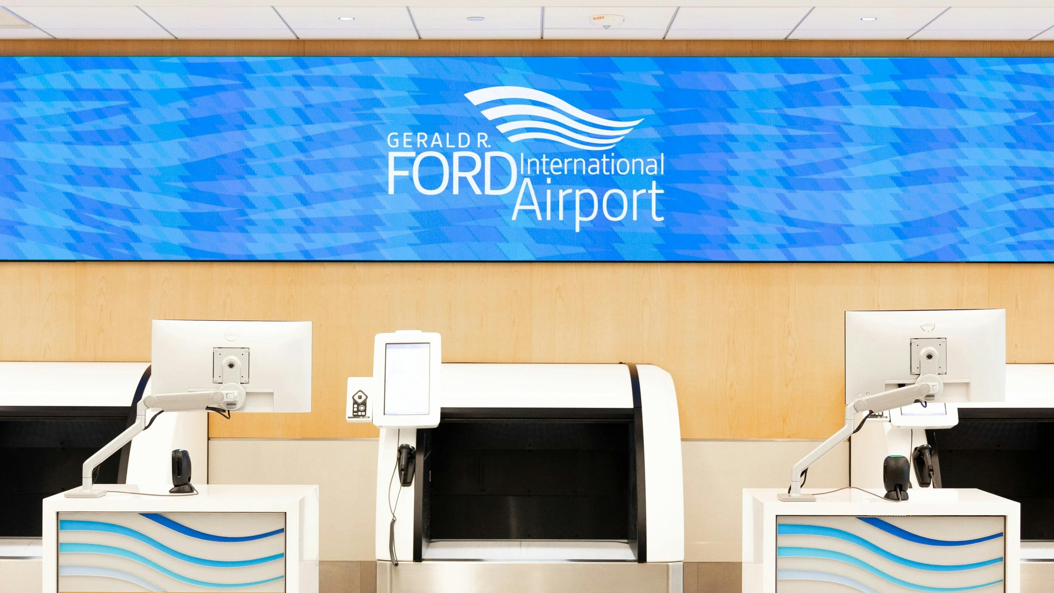 Self-service check-in kiosks and automated bag drop unit at Gerald R. Ford International Airport, with airport branding displayed on an overhead digital sign.