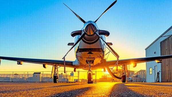 Single-engine turboprop aircraft parked nose-out on the tarmac at sunrise, with propeller blades crossed and a hangar visible in the background.