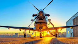 Single-engine turboprop aircraft parked nose-out on the tarmac at sunrise, with propeller blades crossed and a hangar visible in the background.