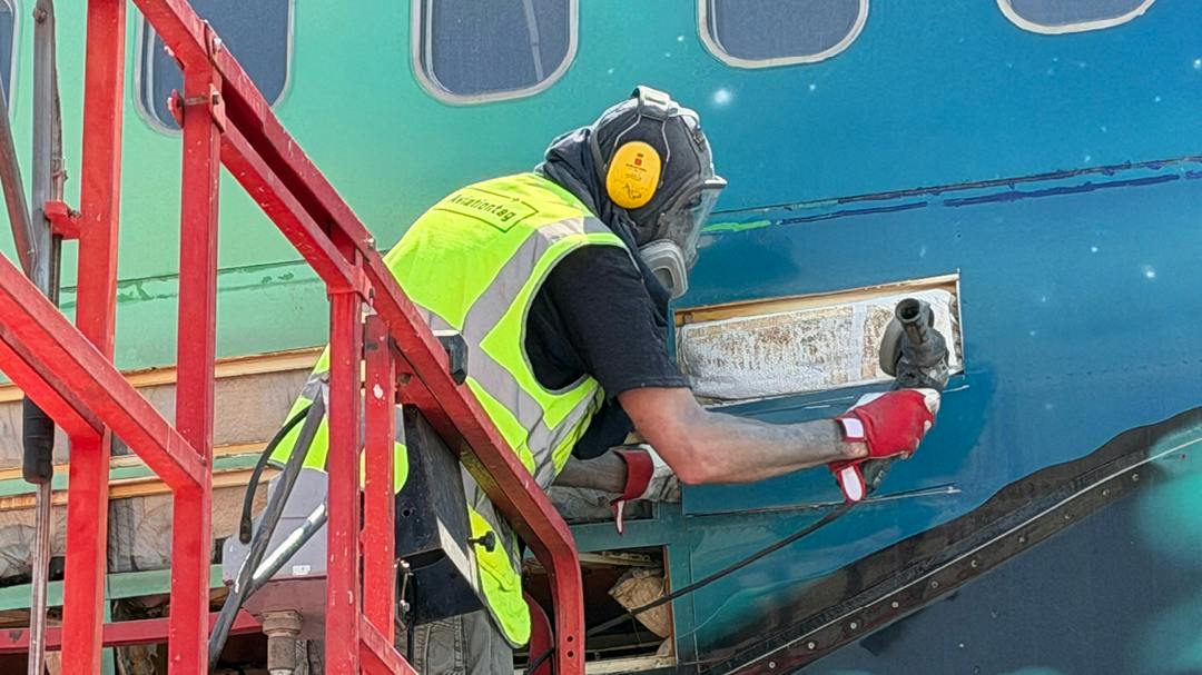 A technician wearing PPE removing a panel of siding from a blue-and-green airplane