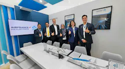 Six people dressed in business attire giving a thumbs-up to the camera while standing behind a table showcasing model airplanes and documents