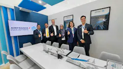 Six people dressed in business attire giving a thumbs-up to the camera while standing behind a table showcasing model airplanes and documents