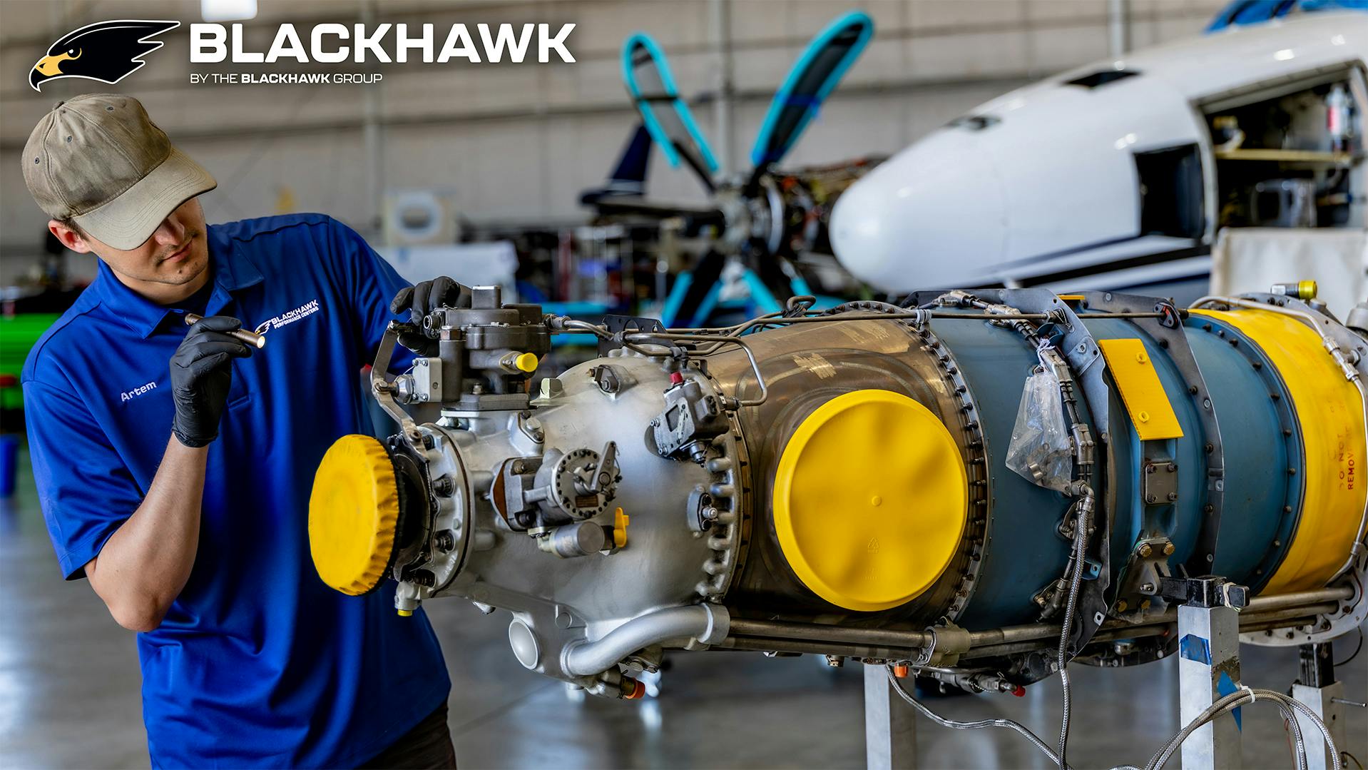 A technician wearing a blue polo working on an airplane engine, with the word 'BLACKHAWK' printed at the top of the photograph